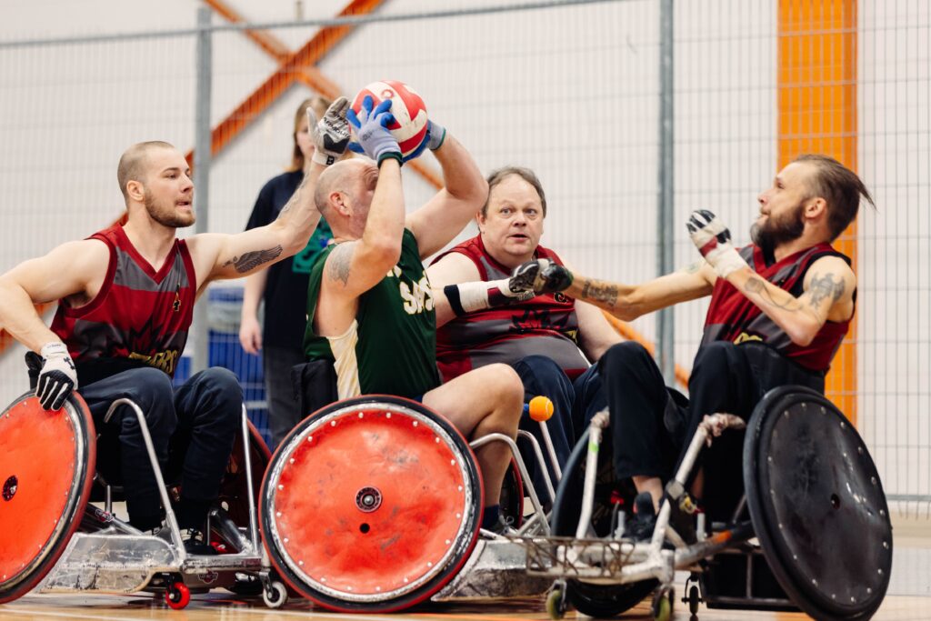 Four wheelchair rugby players are in action, with one closest to the camera about to throw the ball.